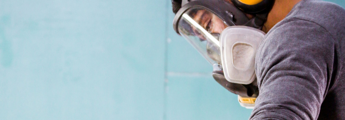 Worker wearing a full-face reusable respirator and hearing protection while using a handheld power tool in a dusty workspace.