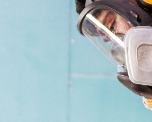 Worker wearing a full-face reusable respirator and hearing protection while using a handheld power tool in a dusty workspace.