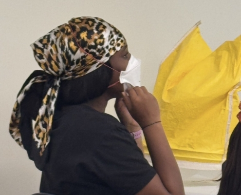 Student wearing a white mask with hands near the chin during a MaskFit+ session, with yellow fit-testing hoods nearby.