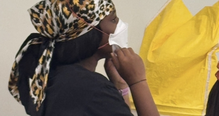 Student wearing a white mask with hands near the chin during a MaskFit+ session, with yellow fit-testing hoods nearby.