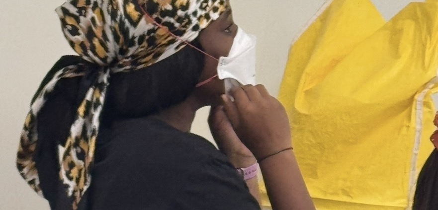 Student wearing a white mask with hands near the chin during a MaskFit+ session, with yellow fit-testing hoods nearby.